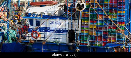 Ein blaues Fischerboot mit bunten Kisten liegt an einem belebten Hafen an. Essaouira, Marokko Stockfoto