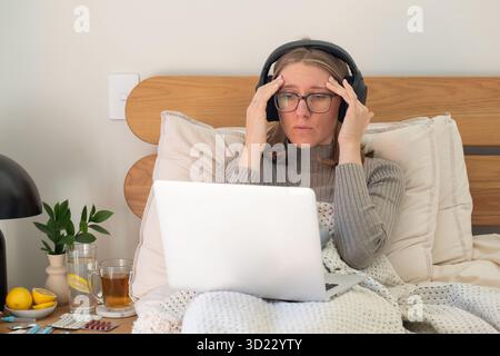 Junge Frau mit Kopfschmerzen, die im Bett auf dem Laptop arbeitet. Fernarbeit, Krankheit. Stockfoto