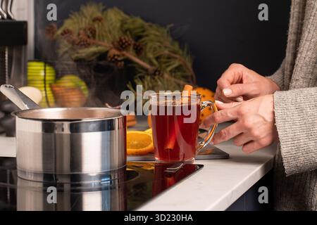 Eine Frau steht neben dem Herd mit einem Topf Glühwein und hält ein Glas Glühwein. Stockfoto
