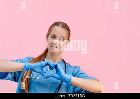 Junge kaukasische Ärztin in blauer Uniform auf einem isolierten rosa Hintergrund zeigt ein Herz mit Handschuhen, einen Ort für Text Stockfoto