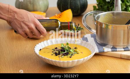 Weibliche Hand, die Olivenöl in Kürbissuppe mit Mikrogrün auf Holztisch gießt Stockfoto