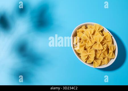 Ungekochte Pasta mit Fliege, Farfalle in einer weißen Schüssel auf blauem Hintergrund mit Schatten Stockfoto