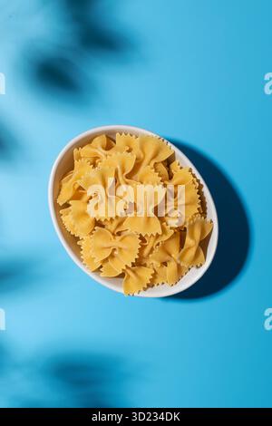 Ungekochte Pasta mit Fliege, Farfalle in einer weißen Schüssel auf blauem Hintergrund mit Schatten Stockfoto