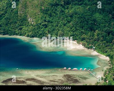 Blick aus der Vogelperspektive auf eine üppig grüne Dschungelküste mit türkisfarbenem Wasser und Bungalows über dem Wasser. Saleman Village, Seram Island, Indonesien Stockfoto