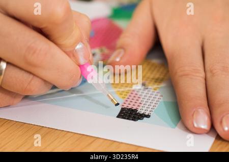 Eine Nahaufnahme einer Hand, die Kristalle auf einer Leinwand für ein Diamantmalprojekt platziert. Diamantmosaik Stockfoto