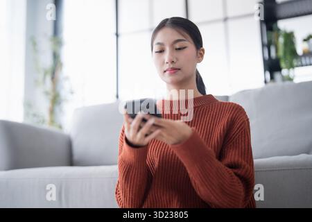 Asiatische Frau, die Smart Home kontrolliert. Mobiltelefon mit Smart-Home-App im Wohnzimmer. Touchscreen-Ai-Technologie. Fernbedienungsanschluss Stockfoto
