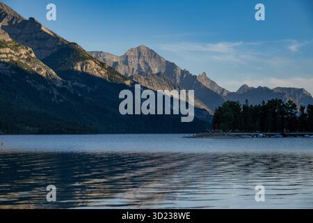 Ruhiger Bergsee mit einer Kulisse aus zerklüfteten Gipfeln bei Sonnenaufgang unter einem klaren blauen Himmel. Waterton Park, Alberta, Kanada Stockfoto