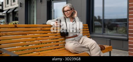 Frau, die auf einer Holzbank vor dem modernen Gebäude sitzt, während sie an bewölkten Tagen ihr Smartphone benutzt Stockfoto