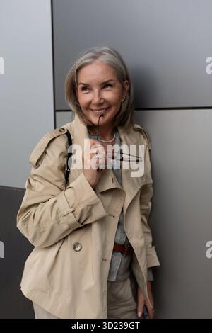 Eine ältere Frau lächelt selbstbewusst und hält eine Brille in einem modernen, urbanen Ambiente während der Tageslichtstunden in einem stilvollen Look Stockfoto