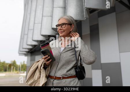 Reife Frau, die Kaffee und Telefon hält, während sie in der Nähe moderner Architektur in einer urbanen Umgebung steht Stockfoto