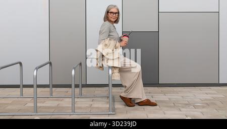 Eine ältere Frau, die tagsüber zwanglos auf einem Fahrradträger sitzt und ein Getränk in der Nähe des modernen Gebäudes hält Stockfoto