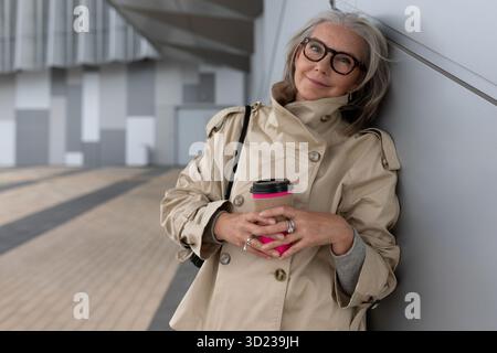 Stilvolle Frau genießt tagsüber Kaffee, während sie sich in einer städtischen Umgebung an eine Wand lehnt Stockfoto
