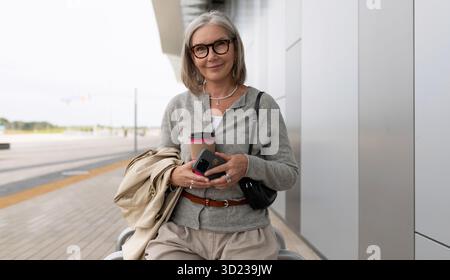 Frau, die an einem bewölkten Tag am Flughafenterminal eine Kaffeetasse und ein Smartphone hält Stockfoto