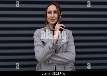 Rothaariger ernsthafter Geschäftsmann mit einem Handy in einem Mantel vor dem Hintergrund einer reliefgrauen Wand Stockfoto