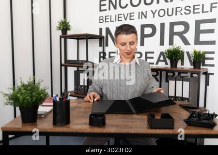 Eine reife Geschäftsfrau mit einem kurzen Haarschnitt auf dem Schreibtisch sieht sich den Ordner mit Dokumenten an Stockfoto