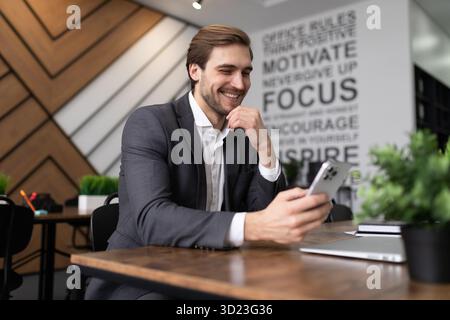 Junger erfolgreicher Geschäftsmann im formellen Anzug im Büro schaut mit einem Lächeln auf den Bildschirm des Telefons Stockfoto