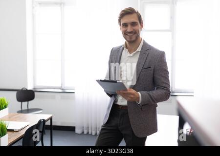 Ein junger Geschäftsmann mit einem elektronischen Tablet in der Hand steht mit einem Lächeln mitten im Büro Stockfoto