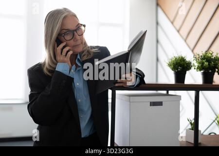 Eine ältere Geschäftsfrau mittleren Alters mit einem Notizblock in der Hand spricht im Büro auf einem Mobiltelefon, Prinzipien und Regeln des Unterrichts Stockfoto