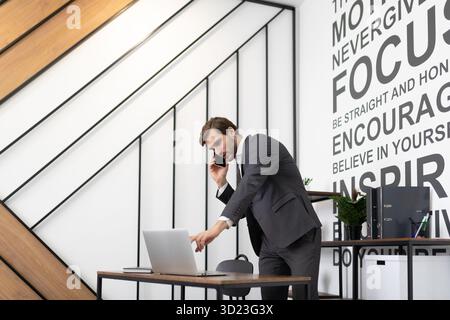 Erwachsener kaukasischer Geschäftsmann im Business-Anzug, der am Telefon spricht und im Büro auf den Laptop-Bildschirm schaut Stockfoto