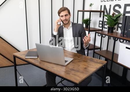 Immobilienmakler, der auf einem Mobiltelefon mit einem Kunden im Büro vor einem Laptop spricht Stockfoto