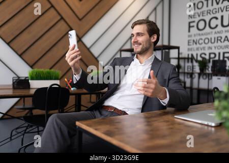 Ein erfolgreicher Geschäftsmann in einem Business-Anzug kommuniziert per Smartphone-Video-Link, während er am Tisch im Büro sitzt Stockfoto