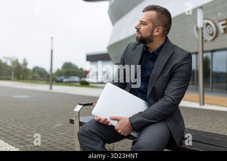 Porträt eines erfolgreichen, reiferen Geschäftsmannes mit Laptop, der neben dem Bürogebäude sitzt Stockfoto
