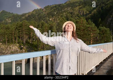 Fröhliche kaukasische junge Frau mit Hut, die die Natur auf der Brücke mit Regenbogen feiert Stockfoto