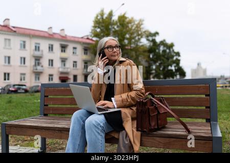 Eine Geschäftsfrau im Park sitzt auf einer Bank und spricht auf einem Mobiltelefon und arbeitet an einem Laptop. Sie arbeitet außerhalb des Bürokonzepts Stockfoto