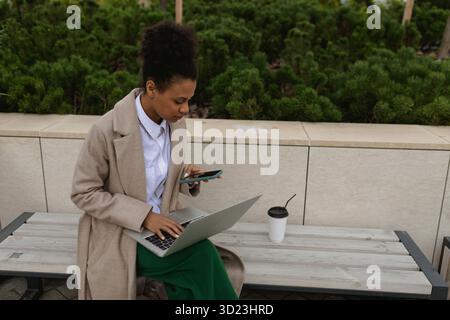 afroamerikanische junge Frau, die an einem Laptop arbeitet und auf den Smartphone-Bildschirm blickt, die im Park neben dem Büro sitzt Stockfoto