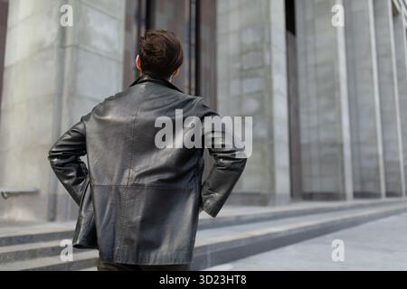 Ein reifer Mann in einer Lederjacke steht und sieht die Gebäude in der Stadt, den Blick von hinten Stockfoto
