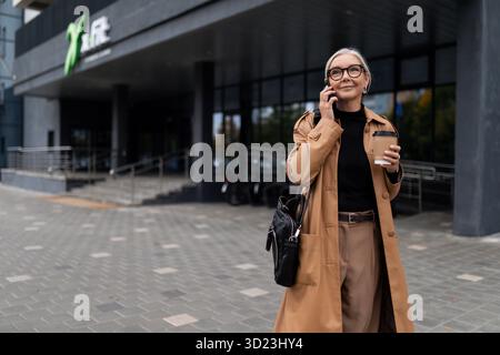 Eine ältere Geschäftsfrau mittleren Alters läuft in der Nähe des Business Centers und spricht über ein Mobiltelefon, Prinzipien und Regeln des Erfolgs Stockfoto