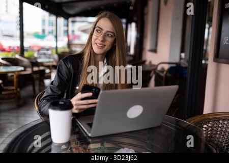 Eine junge Freiberuflerin trinkt Kaffee und arbeitet online an einem Laptop in einem Stadtcafé mit einem Lächeln in die Kamera Stockfoto