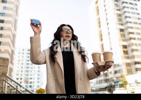 Junge, stilvolle Frau mit zwei Tassen Kaffee in den Händen, die einem Freund in der Stadt winkt Stockfoto
