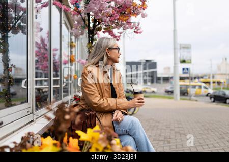 Eine stilvolle Rentnerin sitzt auf einer Bank neben einem Geschäft mit einem Mobiltelefon in der Hand und blickt in die Ferne Stockfoto