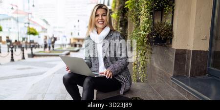 Erwachsene Frau, die außerhalb des Büros am Laptop arbeitet, auf Stufen sitzend, lächelnd in die Kamera schauend, Versicherungskonzept für Geschäftsrisiken Stockfoto