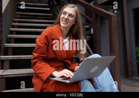 Eine junge Frau, die online an einem Laptop arbeitet und auf einer Treppe neben einem Café sitzt und lächelt und wegblickt Stockfoto