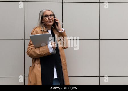 Moderne Geschäftsfrau, die auf einem Mobiltelefon mit einem Laptop in der Hand vor dem Hintergrund einer beigefarbenen Wand spricht Stockfoto