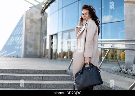 Stilvolle junge Geschäftsfrau am Eingang des Business Centers, die mit einem Mobiltelefon spricht und sich umsieht, stark und unabhängig Stockfoto