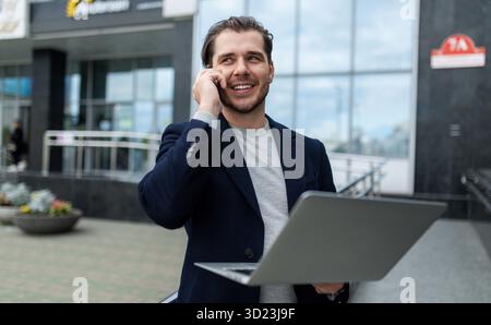 Zufriedener und erfolgreicher Mann, der 35 Jahre alt ist und Geschäftskleidung trägt, spricht am Telefon mit einem Laptop in der Hand Stockfoto