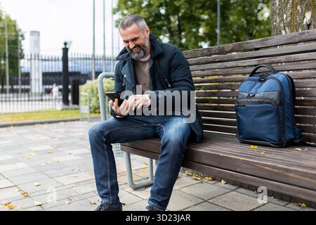 Der fünfzigjährige Tourist, der auf einer Bank im Park mit Rucksack isst, schaut in ein Handy und lächelt Stockfoto