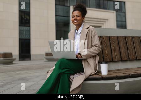 Eine ziemlich erfolgreiche junge afroamerikanerin, die online an einem Laptop arbeitet und auf einer Bank sitzt, mit einem Glas Kaffee auf dem Rücken Stockfoto