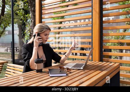 Porträt einer jungen Geschäftsfrau, die am Telefon hinter einem Laptop im Freien spricht Stockfoto