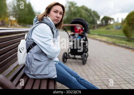 Die verärgerte junge Mutter sitzt im Park auf einer Bank weg vom Kinderwagen mit einem wütenden Gesicht. Das Konzept der postpartalen de Stockfoto