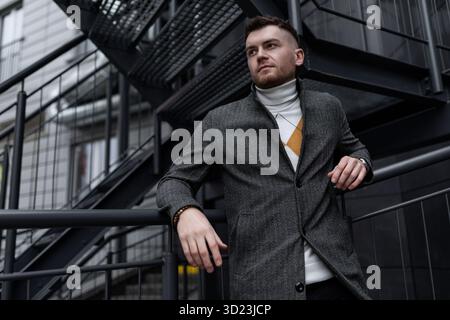 Ein starker junger Mann im grauen Mantel steht auf der Metalltreppe eines trendigen Gebäudes Stockfoto