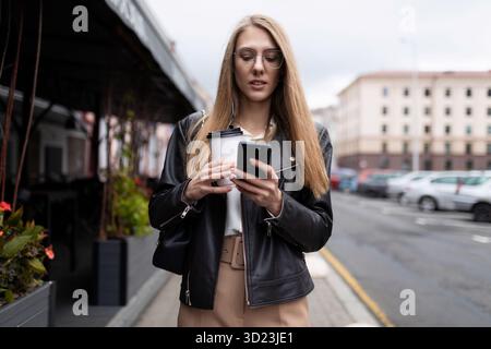 Eine junge Frau, die eine Nachricht auf einem Mobiltelefon mit einer Tasse Kaffee in der Hand vor dem Hintergrund der städtischen Landschaft nex schreibt Stockfoto