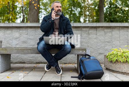 Geschäftsmann in Smart-Casual-Kleidung spricht auf einem Mobiltelefon und arbeitet online in einem Stadtpark an einem Laptop Stockfoto