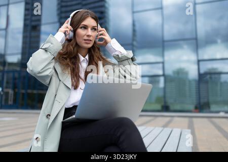 Eine junge Frau in Kopfhörern mit einem Laptop auf den Knien hört Musik vor dem Hintergrund einer verglasten Bürofassade Stockfoto