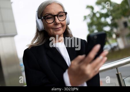 Stilvolle ältere Frau, die mit einem Telefon in der Hand Musik über Kopfhörer hört Stockfoto