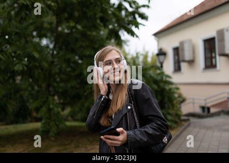 Lächelnde junge Studentin, die Musik mit Kopfhörern vor dem Hintergrund der urbanen Landschaft hört Stockfoto