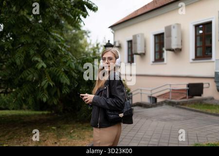 Eine junge Frau läuft durch die Stadt, hört Musik über Kopfhörer und liest Nachrichten auf ihrem Handy Stockfoto
